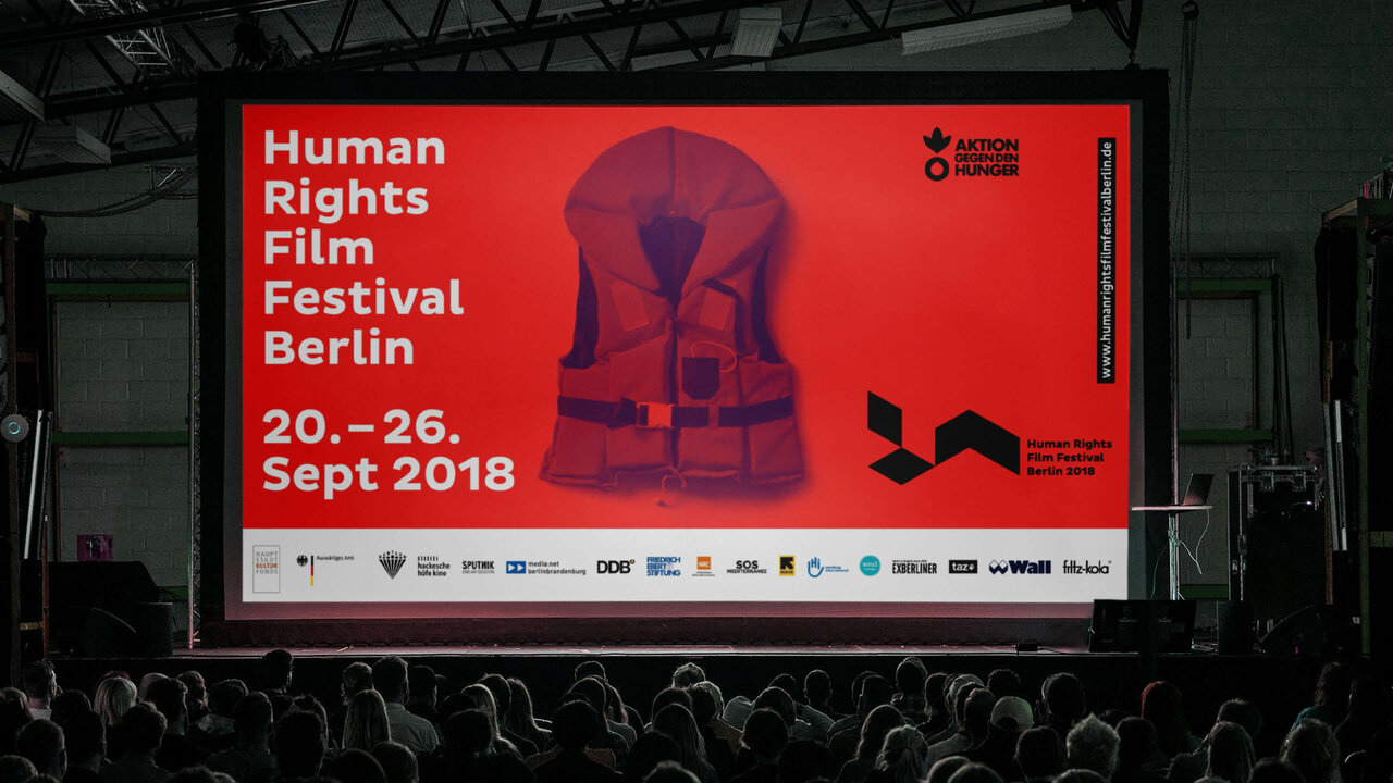 Gro&szlig;es Plakat des Human Rights Film Festivals Berlin mit rotem Hintergrund und stilisierter Rettungsweste, das die Veranstaltung vom 20. bis 26. September 2018 ank&uuml;ndigt, vor einer Zuschauerkulisse in einem Veranstaltungsraum.