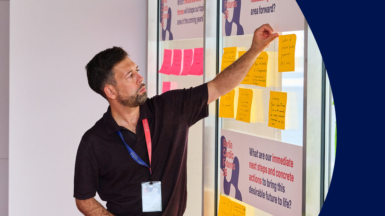 Ein Mann mit Bart und Lanyard steht vor einer Glaswand voller bunter Klebezettel und zeigt auf eine Notiz. Die Wand ist mit Fragen zu n&auml;chsten Schritten und konkreten Ma&szlig;nahmen beschriftet, im Rahmen eines Workshops zur Zukunftsgestaltung.