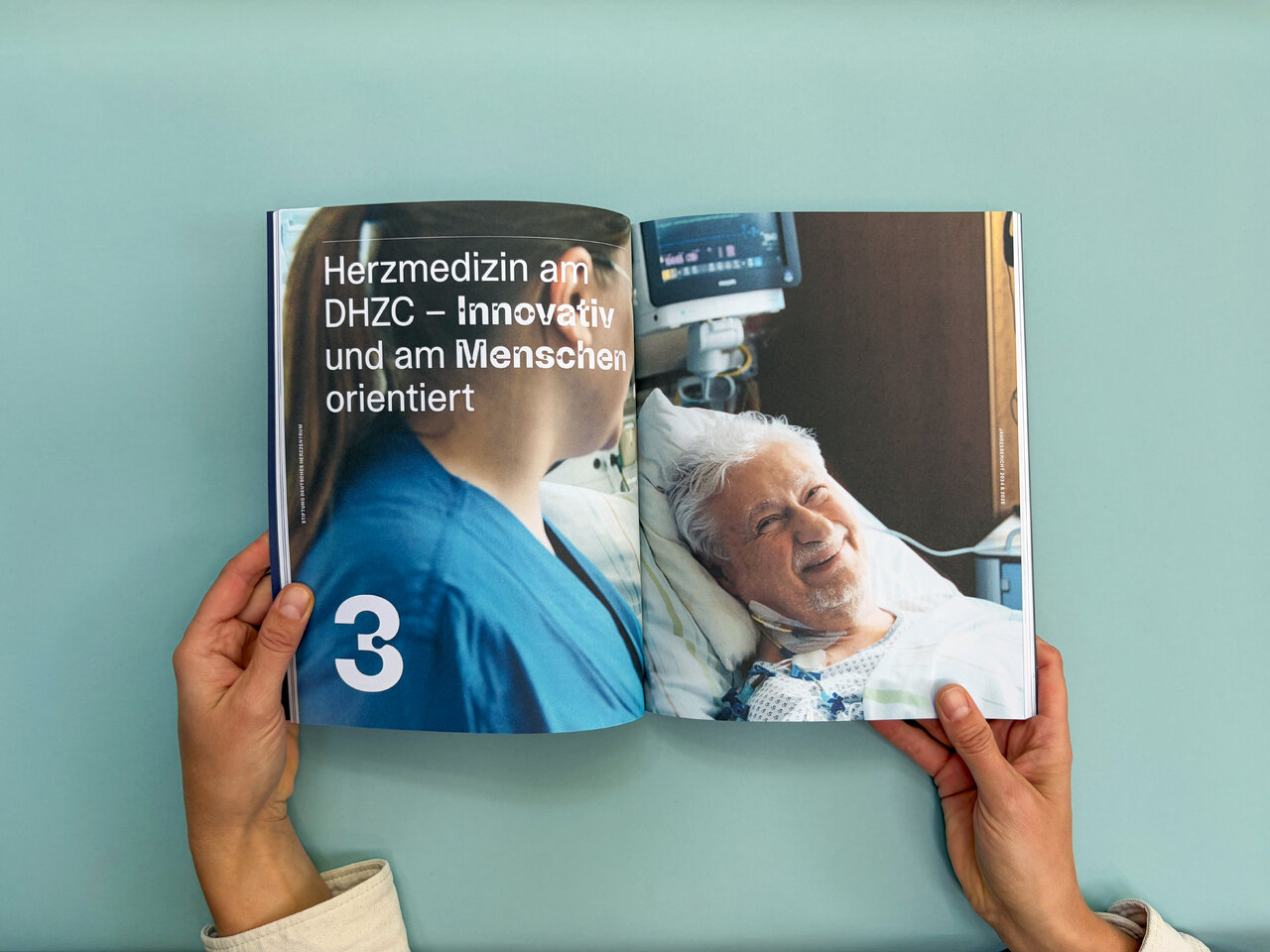 Zwei H&auml;nde halten ein aufgeschlagenes Buch &uuml;ber einem hellblauen Hintergrund. Die linke Seite zeigt den Text &bdquo;Herzmedizin am DHZC &ndash; innovativ und am Menschen orientiert&ldquo; mit der Kapitelnummer 3, die rechte Seite ein Foto eines l&auml;chelnden &auml;lteren Patienten im Krankenbett.