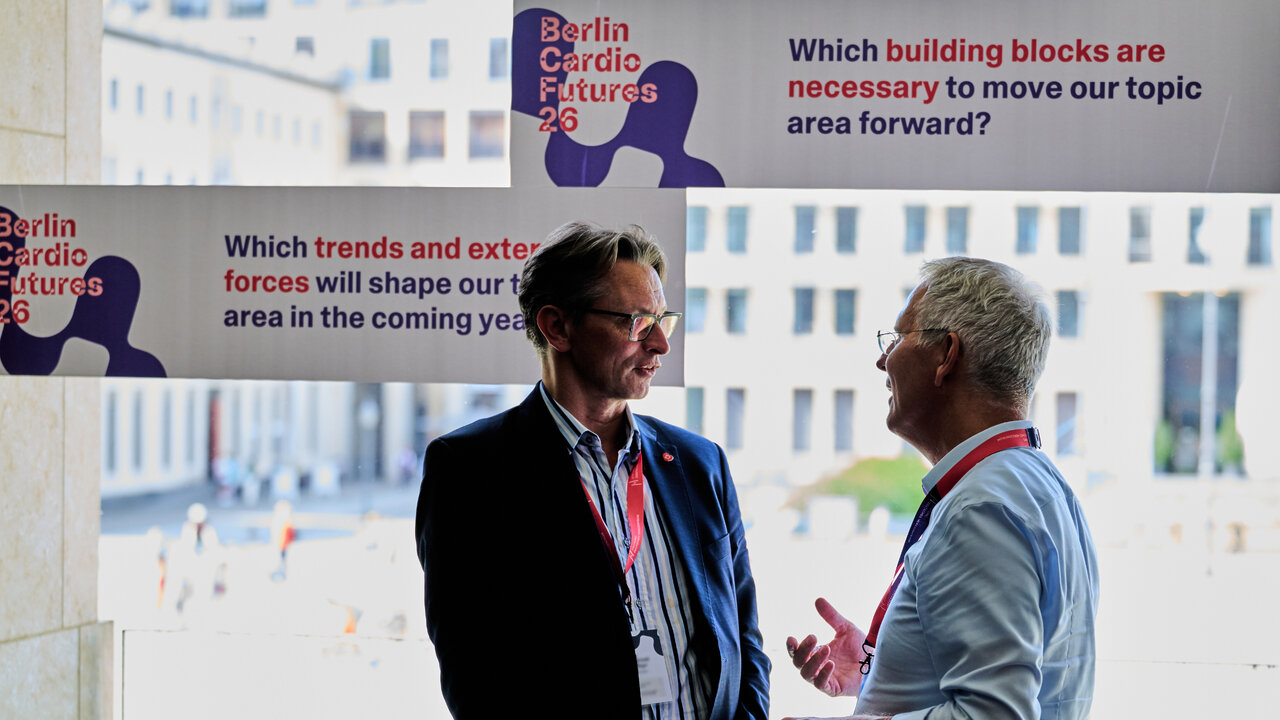 Zwei M&auml;nner mit Konferenz-Namensschildern unterhalten sich vor gro&szlig;en Bannern der Veranstaltung "Berlin Cardio Futures 26" mit Fragen zu Trends und Handlungsfeldern im Gesundheitsbereich. Im Hintergrund ist durch gro&szlig;e Fenster eine Stadtansicht zu erkennen.