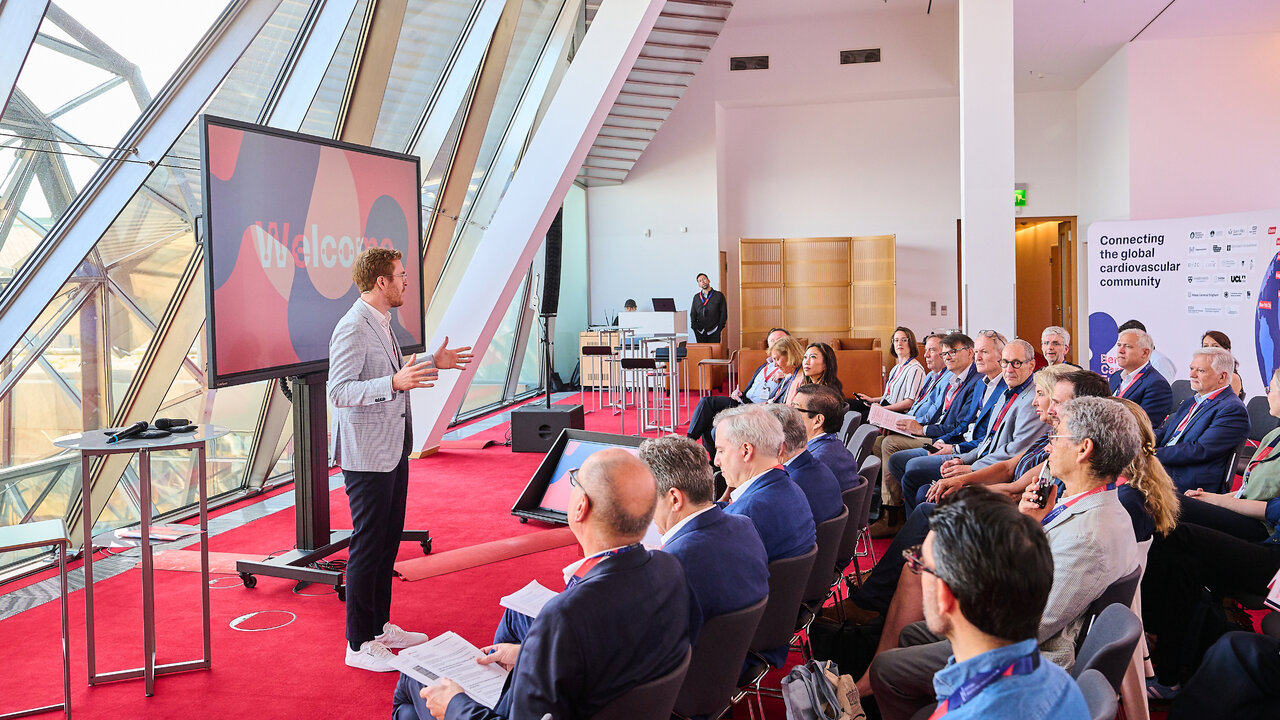 Ein Redner steht vor einem gro&szlig;en Bildschirm mit der Aufschrift &bdquo;Welcome&ldquo; und spricht zu einem sitzenden Publikum in einem modernen Konferenzraum mit schr&auml;gen Glasfenstern. Ein roter Teppich f&uuml;hrt durch den Raum, im Hintergrund ist ein Banner mit dem Text &bdquo;Connecting the global cardiovascular community&ldquo; zu sehen.