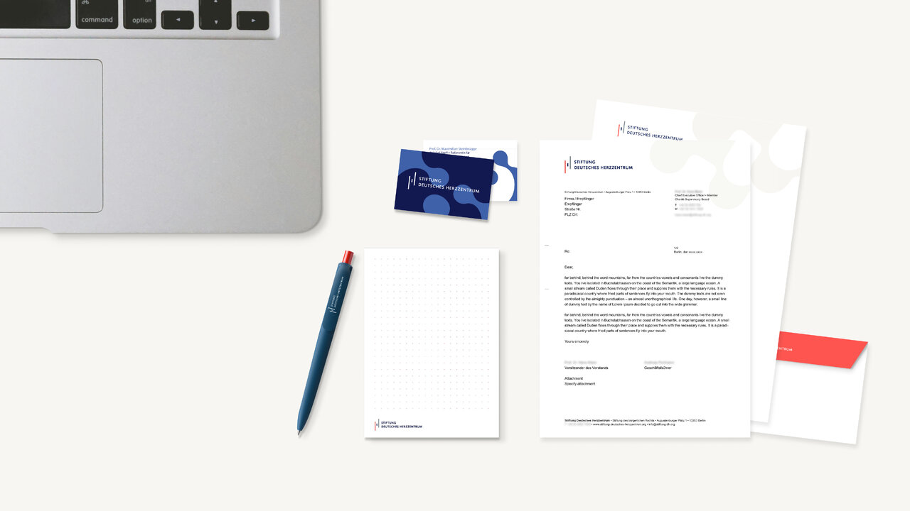 Laptop, blaue Visitenkarten, Briefpapier mit Briefkopf, ein Kugelschreiber und ein roter Umschlag liegen auf einem hellen Schreibtisch &ndash; ein Corporate-Design-Set einer Organisation.