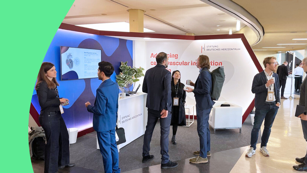 Menschen unterhalten sich an einem Ausstellungsstand mit der Aufschrift &bdquo;Advancing cardiovascular innovation&ldquo; und dem Logo der Stiftung Deutsches Herzzentrum, in einer modernen Eingangshalle mit gebogener Decke.