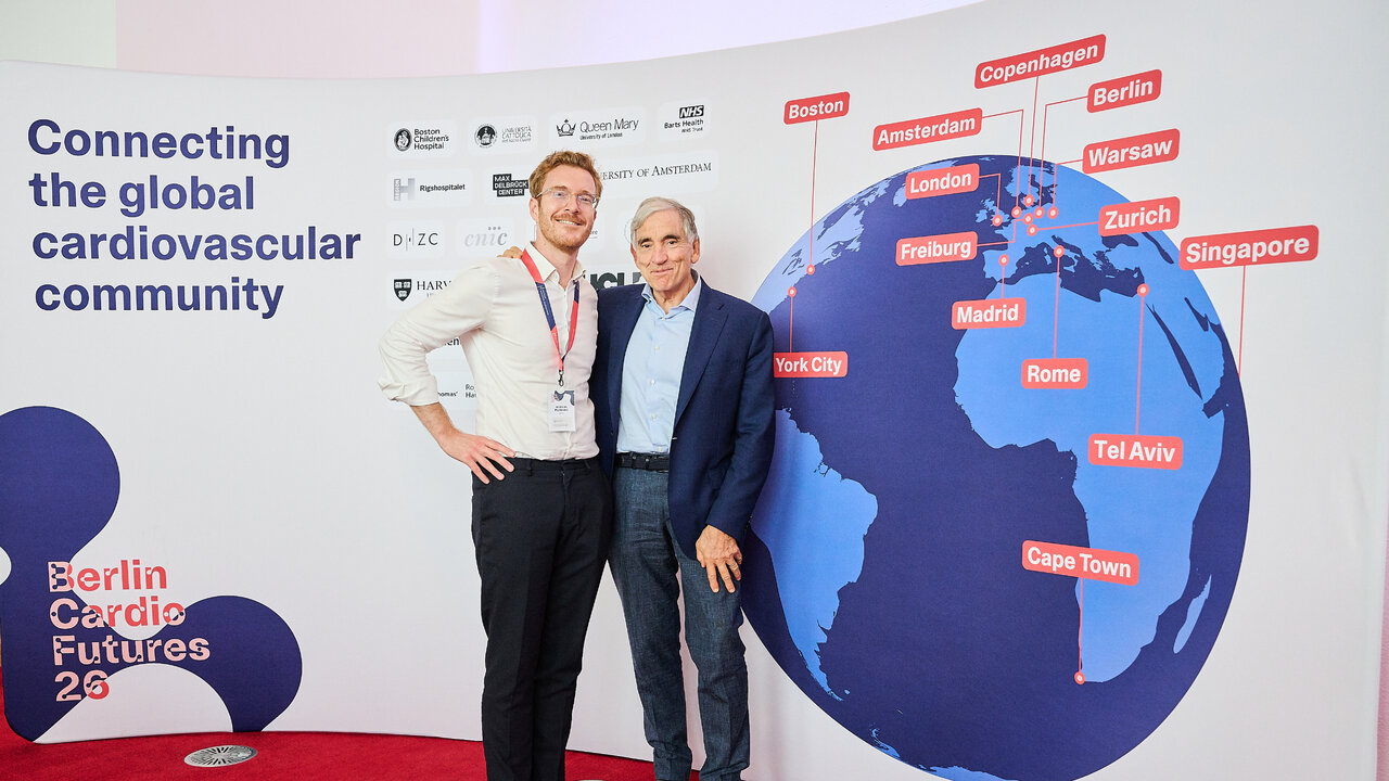 Zwei M&auml;nner stehen l&auml;chelnd vor einer Weltkarten-Grafik mit markierten St&auml;dten wie Berlin, London, Singapur und Kapstadt, die die globale Vernetzung der Kardiologie-Community darstellt. Im Hintergrund ist der Titel &bdquo;Connecting the global cardiovascular community&ldquo; sowie das Logo &bdquo;Berlin Cardio Futures 24&ldquo; zu sehen.
