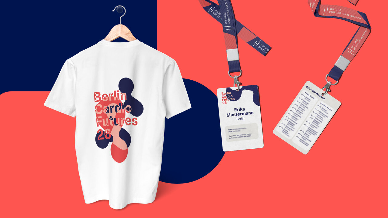 Wei&szlig;es T-Shirt mit abstraktem Grafikmuster in Marineblau und Korallrot sowie dem Schriftzug "Berlin Circle Futures 26", pr&auml;sentiert auf einem Kleiderb&uuml;gel. Daneben h&auml;ngen zwei Namensschilder an gestreiften Lanyards mit Veranstaltungsdetails und Teilnehmerinformationen, vor einem zweifarbigen Hintergrund in Korallrot und Dunkelblau.